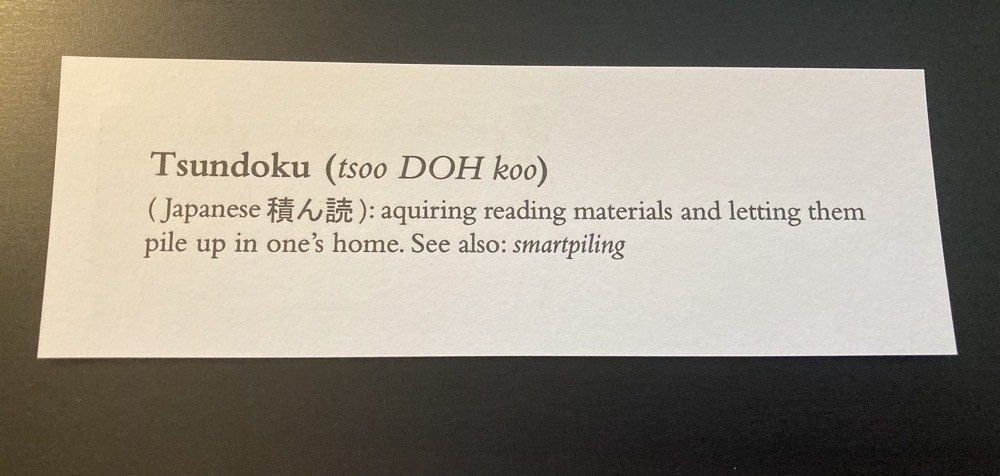 the flipside of the bookmark showing the definition of the word tsundoku: acquiring reading materials and letting them pile up in one's home. See also smartpiling