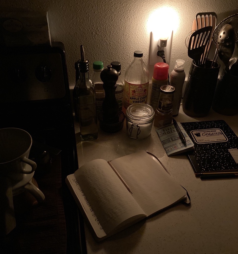 a corner of a kitchen counter in the dark, lit by a nightlight with jars and bottles lined up beside several tall containers holding utensils; three notebooks, two closed and one open to a blank page